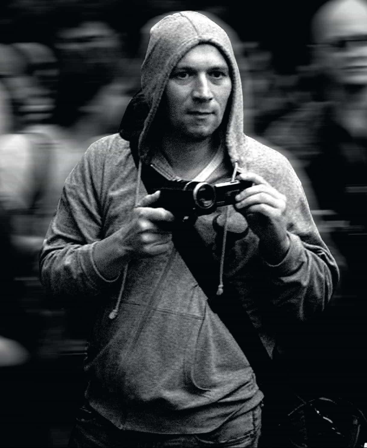 Чёрно-белая фотография мужчины в толстовке с капюшоном, который держит видеокамеру на уровне груди. Он смотрит прямо перед собой. Фон размыт, что создаёт ощущение движения.