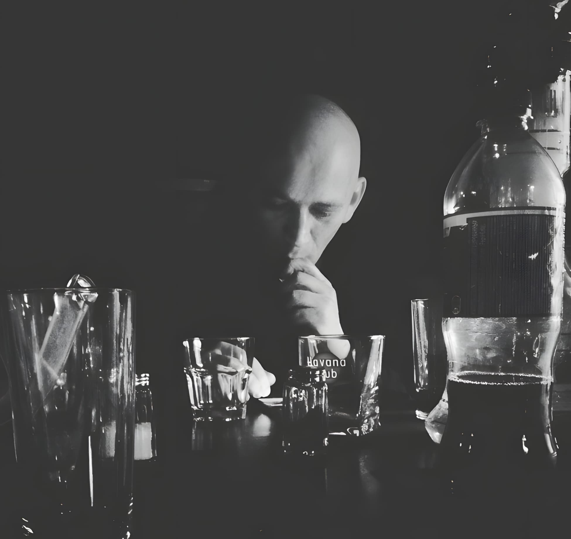 Малоосвещенная черно-белая фотография, на которой на заднем плане смутно освещенный лысый человек смотрит вниз, прикрыв рот рукой. На переднем плане, на поверхности, похожей на барную стойку или стол, стоят несколько стаканов, на одном из которых видна надпись «Havana Club», и большая бутылка, частично наполненная темной жидкостью. Общее настроение мрачное или задумчивое.